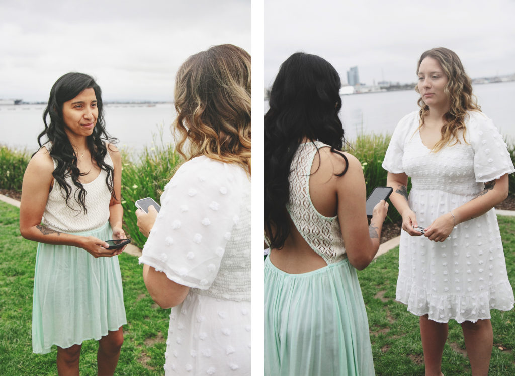At a same-sex wedding ceremony in San Diego, two brides stand intimately, exchanging vows with the calm bay as their backdrop. The first image captures one partner, clad in a mint green dress, holding a smartphone, her eyes glistening with emotion as she smiles tenderly at her soon-to-be wife. In the companion image, the other bride, dressed in a white eyelet dress, holds her own phone, a look of heartfelt sincerity on her face as she listens intently. Their contrasting dresses complement each other, as does their shared gaze, a beautiful testament to their union. The natural setting, with lush grass and reeds swaying gently, creates a serene atmosphere, while the distant cityscape adds an urban contrast to their intimate moment. This diptych tells a story of love, commitment, and the start of a new chapter together under the open skies of San Diego.