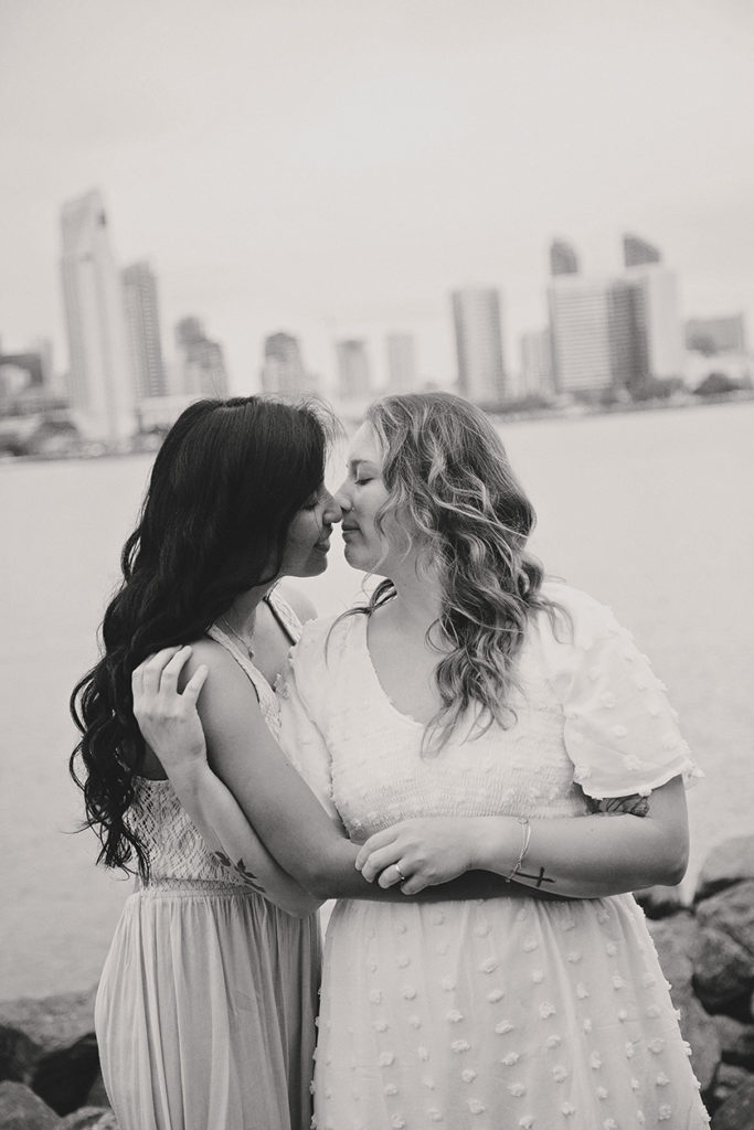 In an intimate monochrome portrait, two brides share a close embrace on their wedding day. Their foreheads gently touch, eyes closed, savoring the moment of closeness. The brides' contrasting dresses—one in delicate white lace, the other in a graceful pastel—complement each other perfectly, just like the connection they share. Behind them, the San Diego skyline fades into a soft blur, the city's presence quiet against the tender scene. This photograph captures the essence of a promise, a whisper of forever held between them, framed by the timeless grace of black and white.