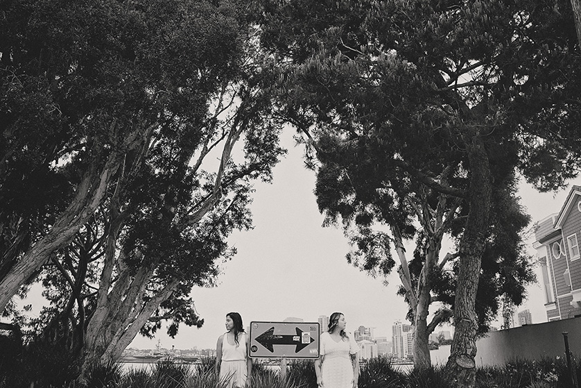 Framed by the whispering eucalyptus, two brides stand apart yet connected, each holding one end of a symbolically crafted sign with a directional arrow, indicating the unity of their paths ahead. The black and white tones lend a classic feel to the urban setting glimpsed in the distance, a city that buzzes beyond their tranquil haven.