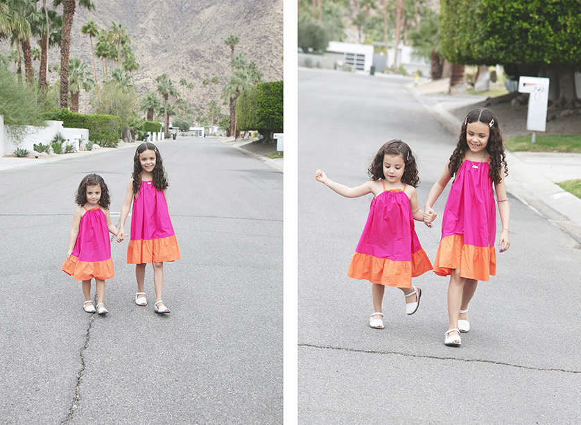 Young sisters in vibrant orange and magenta dresses hold hands and walk down the street