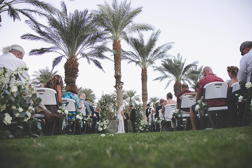 Wide photo of the whole ceremony site, down the aisle with the couple and the alter in the distance, frame by tall palm trees