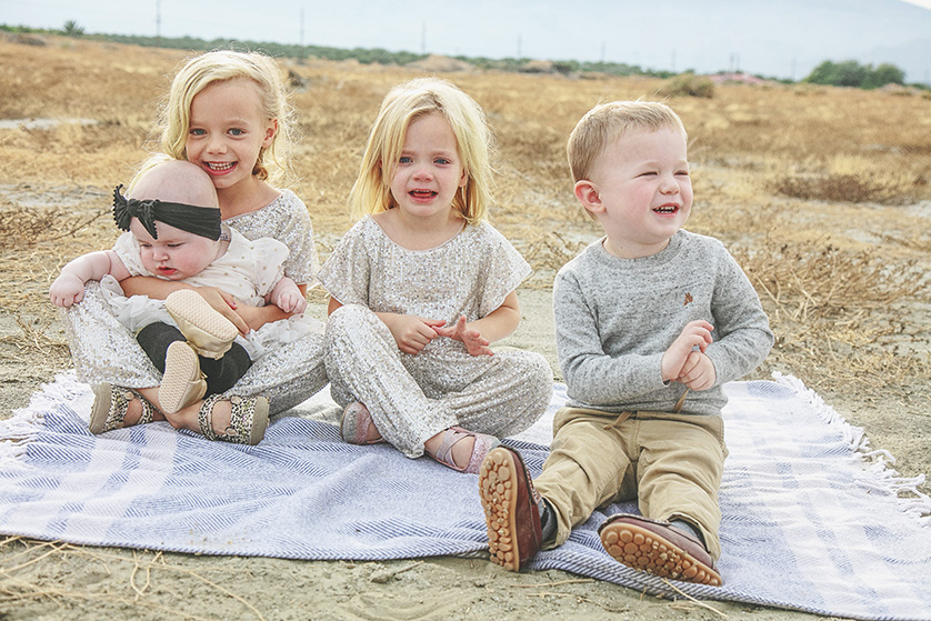 Four young children sitting on a blanket in the sandy desert.