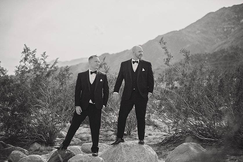 Two grooms wearing black tuxedos and bowties stand on rocks in the Palm Springs area desert