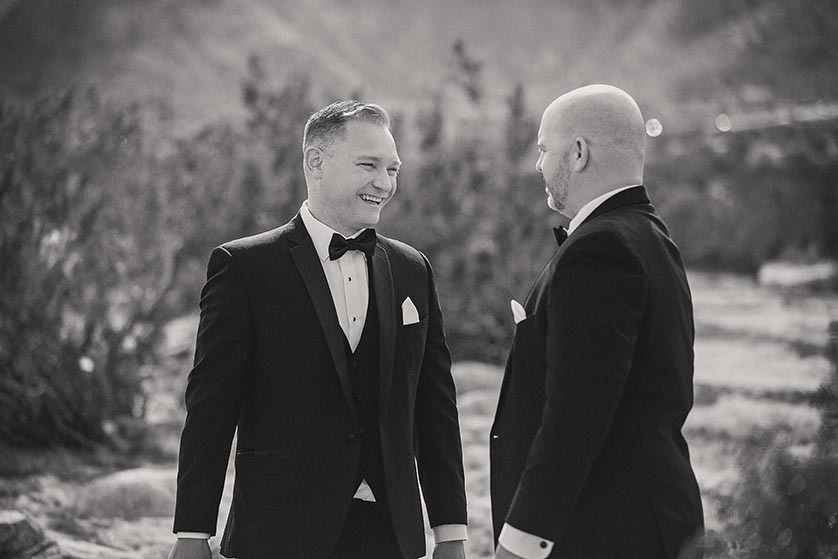 Two grooms laugh and smile in their crisp tuxes in the desert