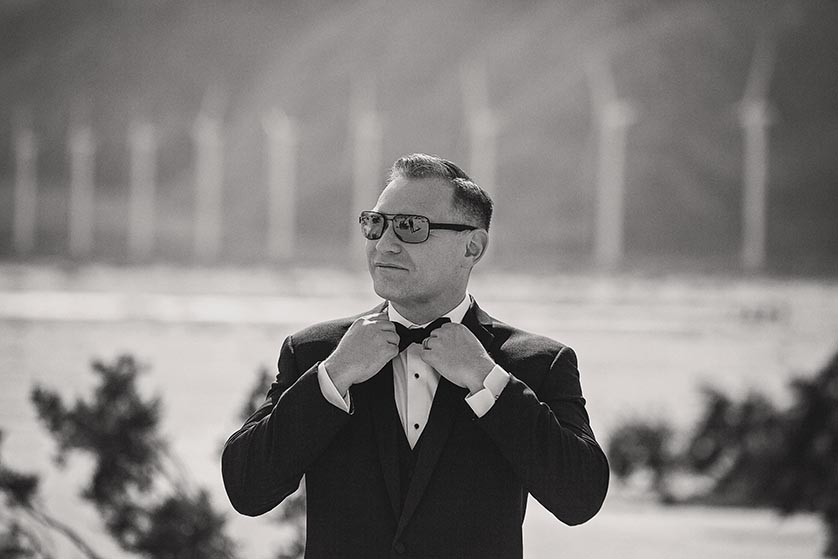 An image of a groom in mirrored sunglasses adjusting his bowtie, a reflection of his husband in the glasses