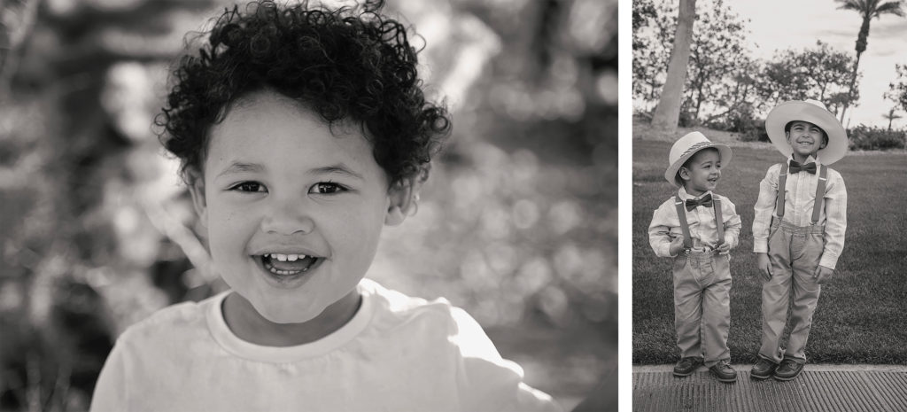 Black and white photo of a curly haired toddler smiling directly at the camera and two young boys in suspenders and hats