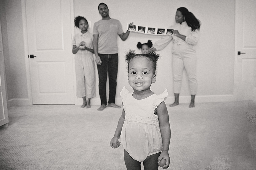 A toddler hams for the camera while her family in the background get posed.