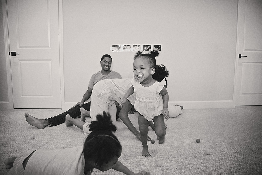 This lively black-and-white photograph captures the essence of family joy and playful chaos. The central figure is a gleeful toddler, her face lit up with an exuberant smile as she runs towards the camera.