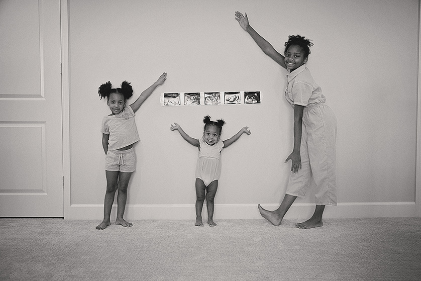 This delightful black-and-white photograph captures three sisters joyfully posing in front of a series of ultrasound photos, marking the anticipation of a new sibling.