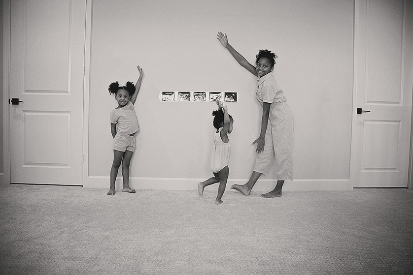 In this charming black-and-white photograph, three sisters joyfully pose in front of a series of ultrasound photos, symbolizing the anticipation of their new sibling.