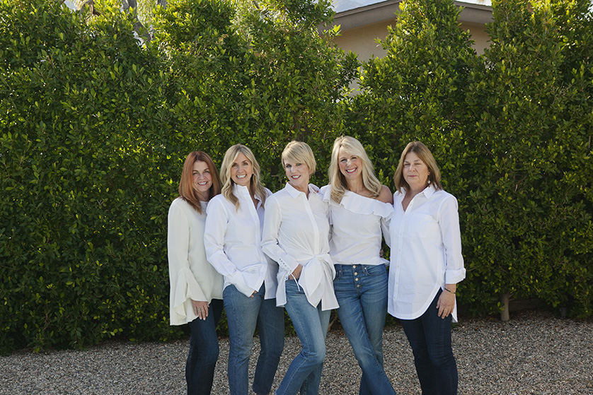 Five smiling women in white shirts and jeans posing outdoors.