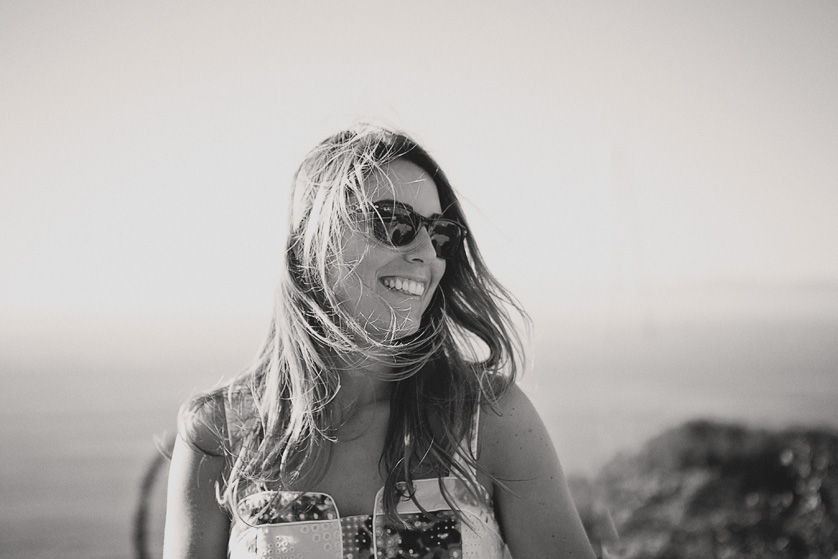 Woman with windswept hair and sunglasses smiles at her fiance