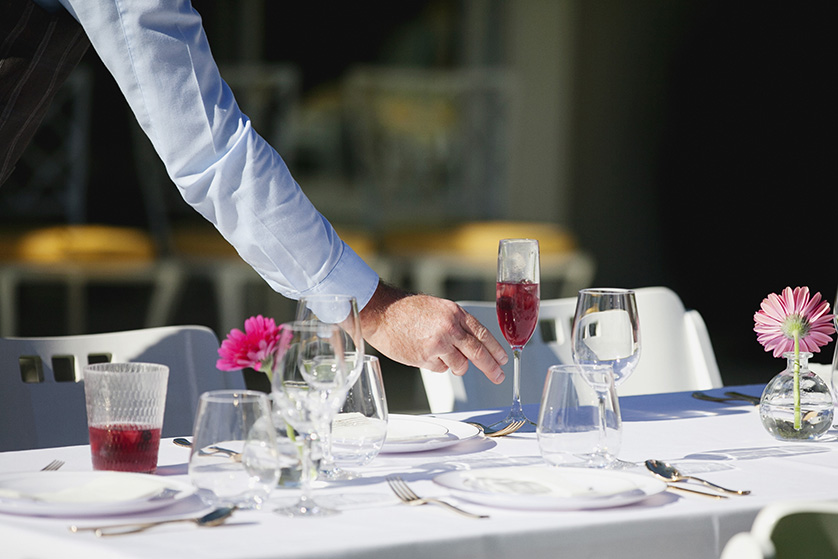 A guest reaches for a champagne flute on a beautifully set wedding table with pink flowers and glassware.