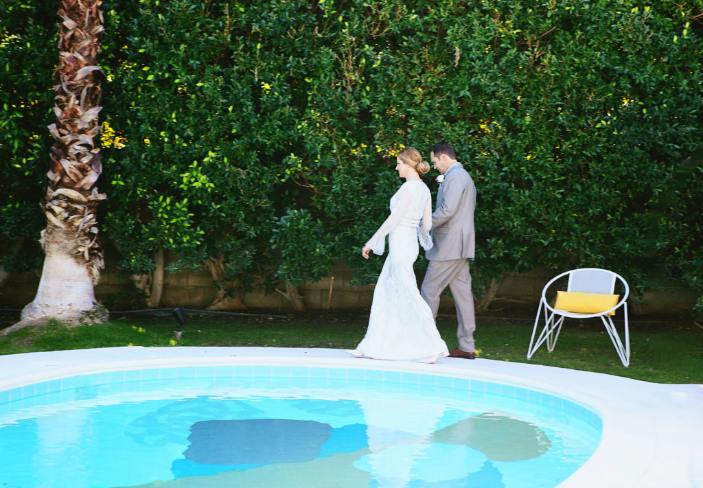 Bride and groom walk hand-in-hand beside a turquoise pool in a lush Palm Springs backyard.