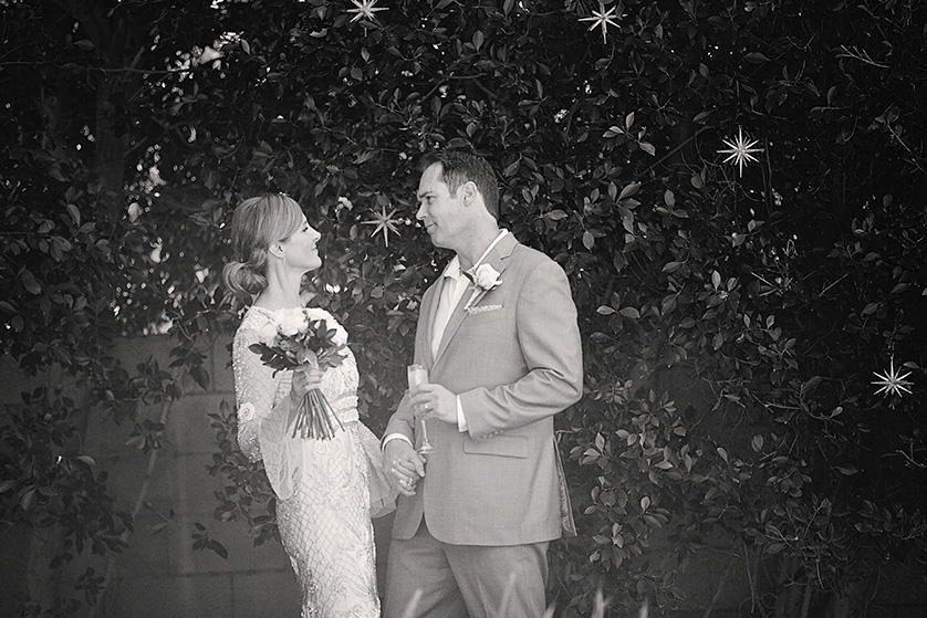 Bride and groom gaze at each other lovingly while holding hands and champagne during their Palm Springs backyard wedding.