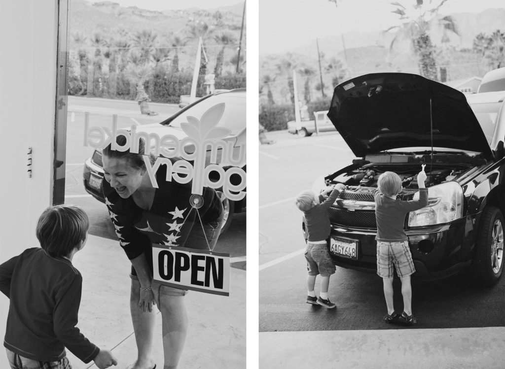 Two young boys pretend to fix a car engine with toy tools outside a Palm Springs shop, while a smiling woman greets them through the glass door.