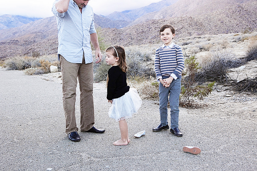 A barefoot little girl in a white tutu stands on a desert road with her father and brother, surrounded by mountains and scattered shoes.