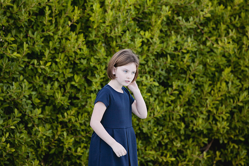 A young girl in a navy blue dress stands pensively in front of a lush green hedge.