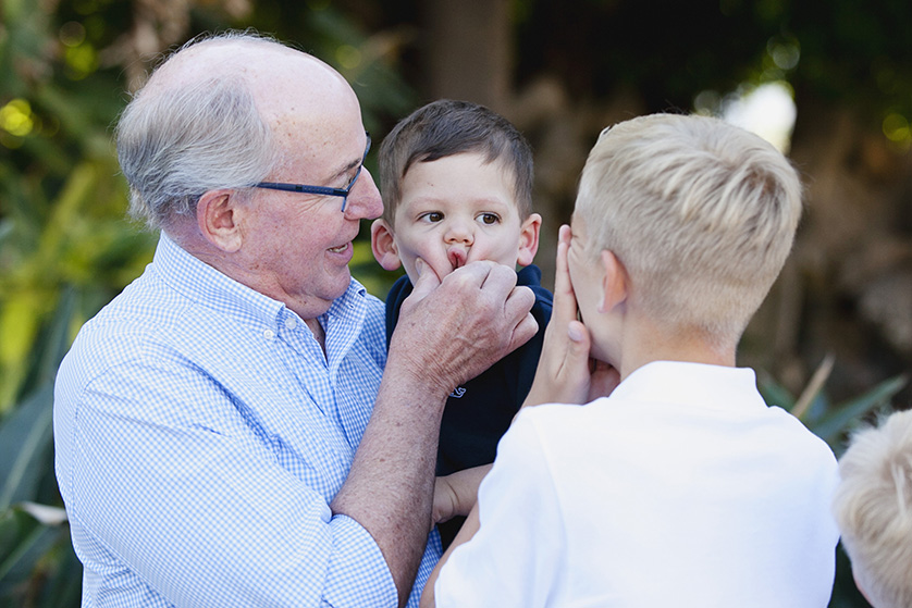 An older man holds a toddler while playfully pinching his nose, surrounded by two young boys sharing a laugh.