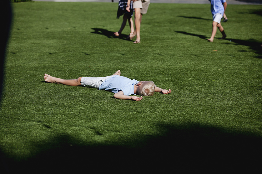 A young boy lies sprawled face-down on a grassy field in the sun, while others walk and play in the background.