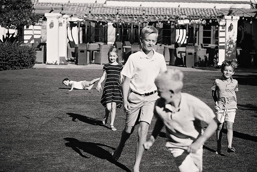 A group of children run and laugh on a sunlit lawn in front of a Spanish-style building, with one child playfully falling behind.