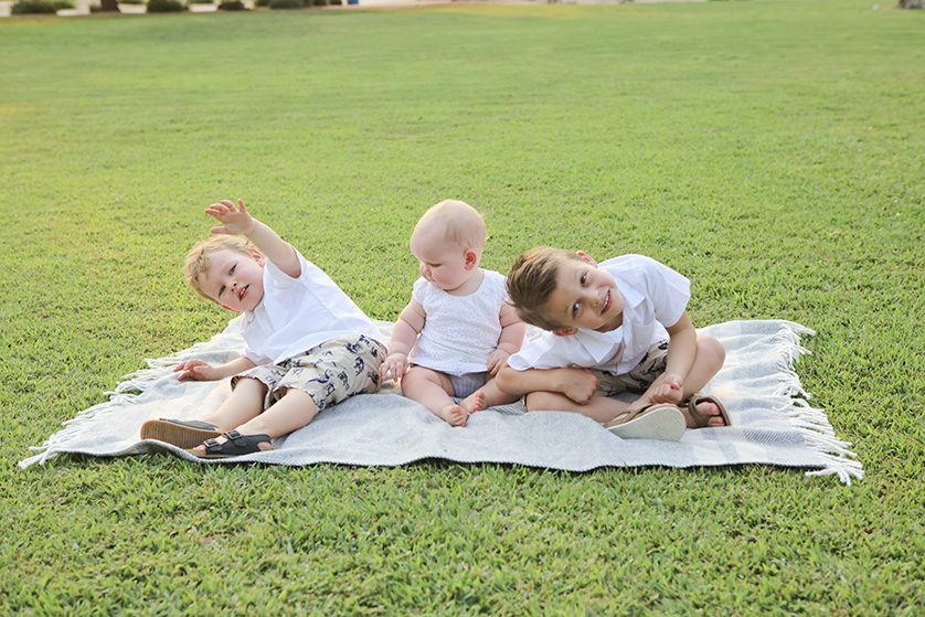 Three young siblings sit on a blanket in the grass, laughing and playing together during a sunny day outdoors.