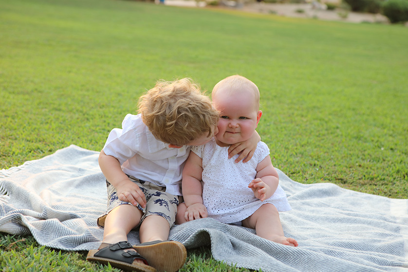 A toddler lovingly cuddles a baby sibling on a blanket in the grass during a warm, sunny day.