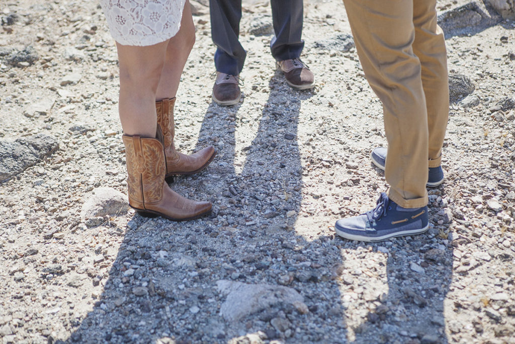 Two brides stand side by side in the desert, one wearing cowboy boots and the other in casual shoes, during their wedding ceremony.