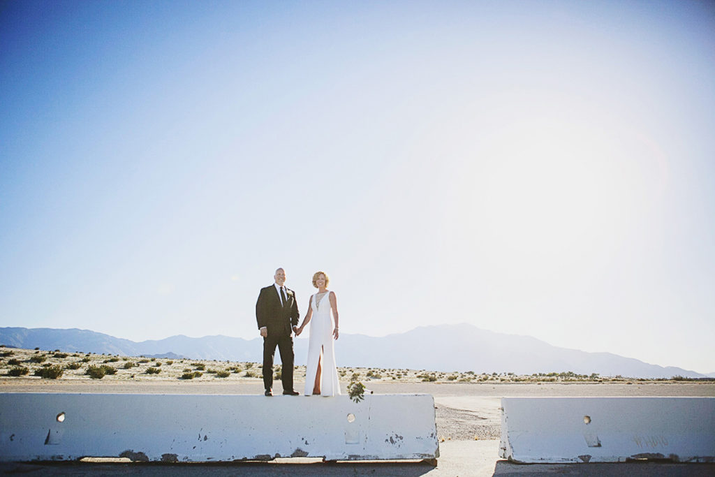 Bride and groom stand hand in hand on a concrete barrier in the desert, with mountains and bright sun in the background.
