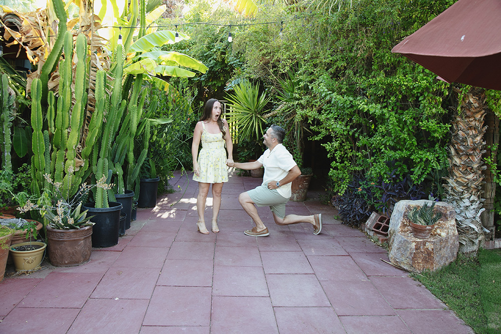 Man proposes on one knee at Casa Monte Vista in Palm Springs as his partner reacts with joyful surprise in a lush garden setting.