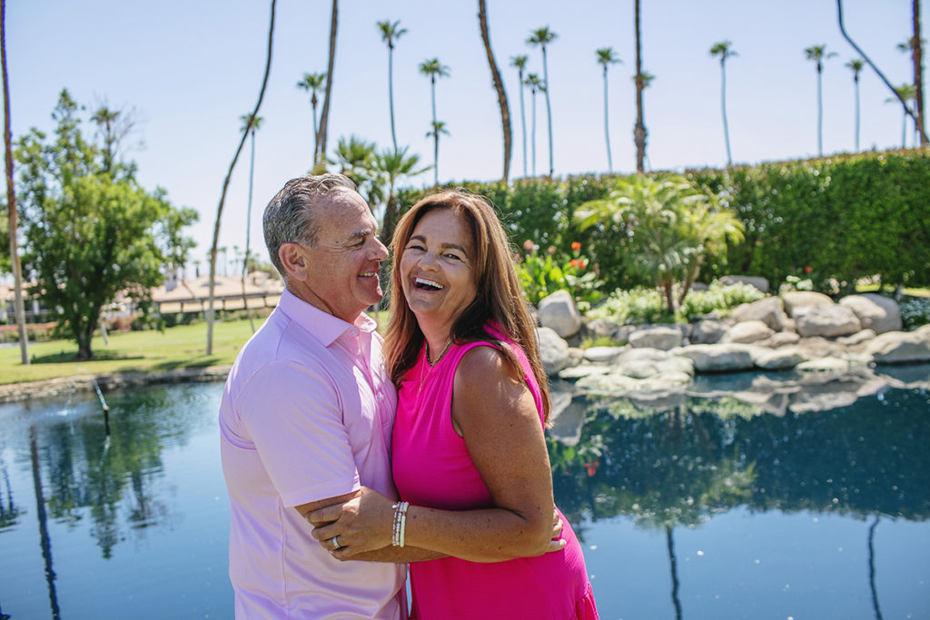 A smiling couple embraces near a reflective pond surrounded by palm trees and lush greenery in Palm Springs.