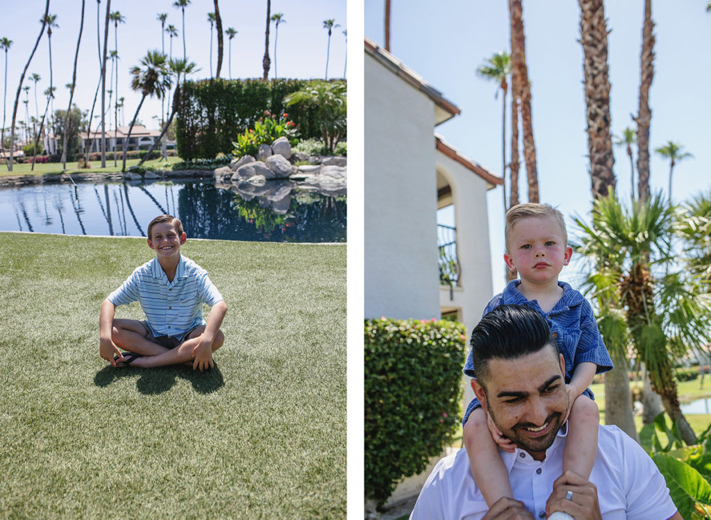 A young boy in a striped shirt sits cross-legged on the grass near a pond with palm trees in Palm Springs.  A father gives his young son a piggyback ride outside a Palm Springs home with palm trees in the background.