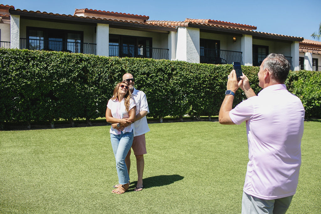 A man takes a phone photo of a smiling couple embracing outdoors at a Palm Springs resort.