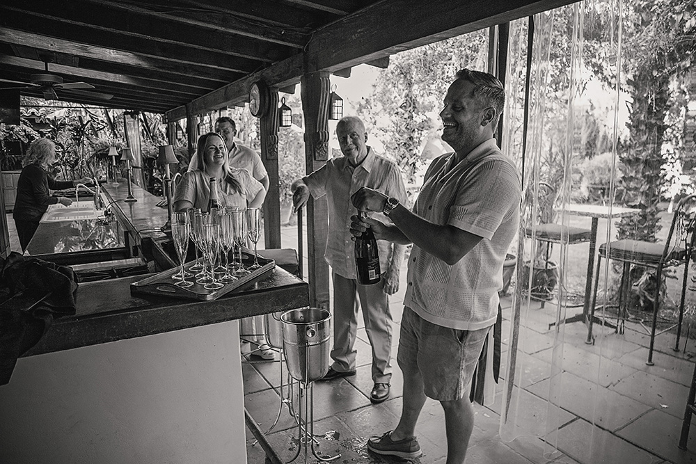 A man opens a bottle of champagne while family and friends smile and gather to celebrate a surprise proposal at Casa de Monte Vista in Palm Springs.