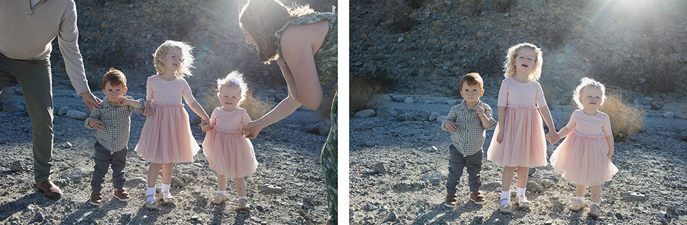 “Three young children standing together during a family photo session in La Quinta, California, with parents just outside the frame offering guidance. The sun glows behind them as they stand on a rocky desert landscape.”