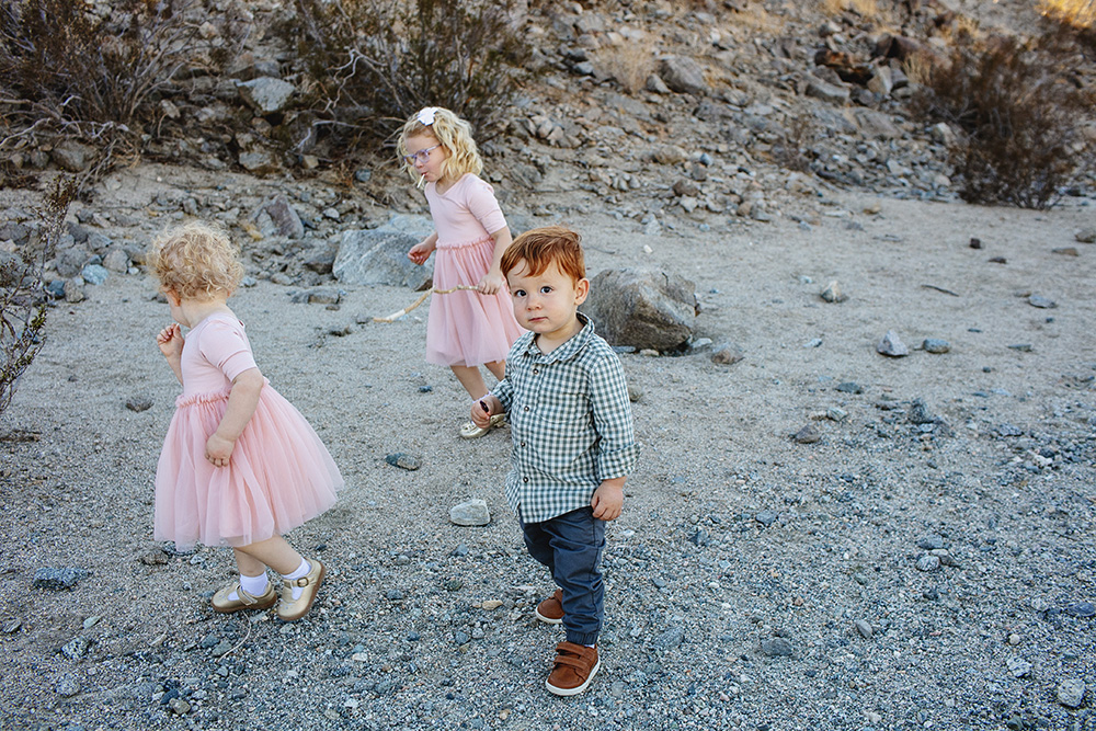 “Three young children exploring a rocky desert area in La Quinta, California. Two little girls in pink dresses walk and spin while a young boy in a checkered shirt looks toward the camera during a candid family photo moment.”