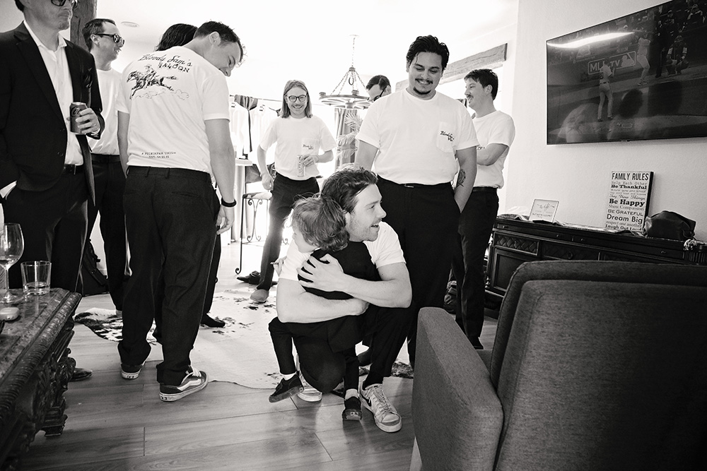 Groom hugging a child while groomsmen gather during getting ready at Bubbling Wells Ranch, captured in black and white documentary wedding photography