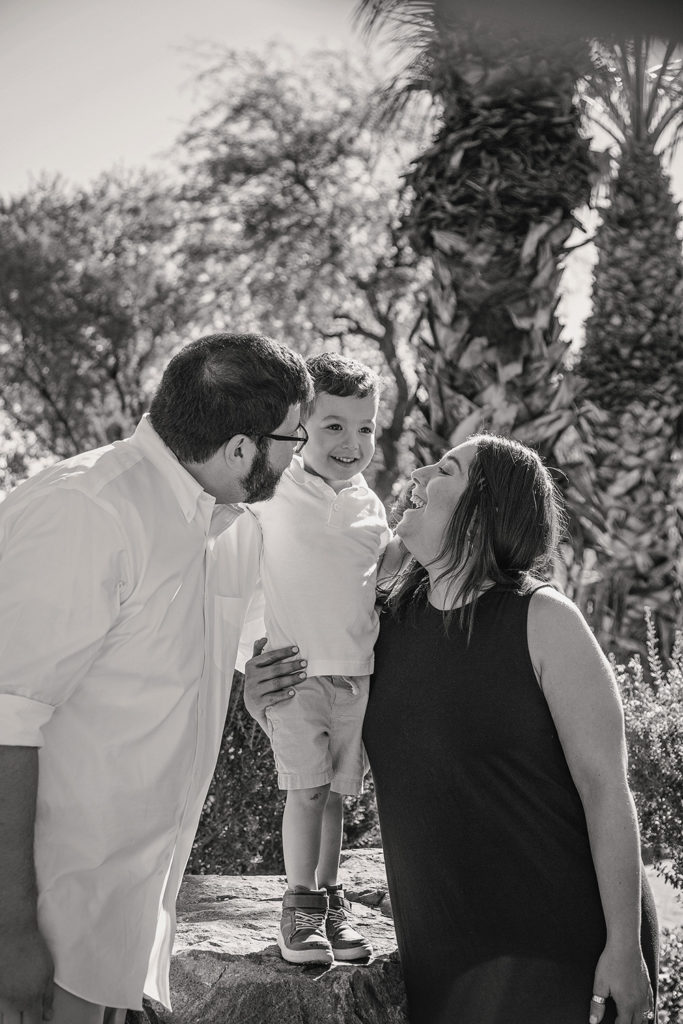 Parents smiling and leaning in toward their young son as he stands on a rock outdoors in Palm Springs, sharing a joyful moment together.