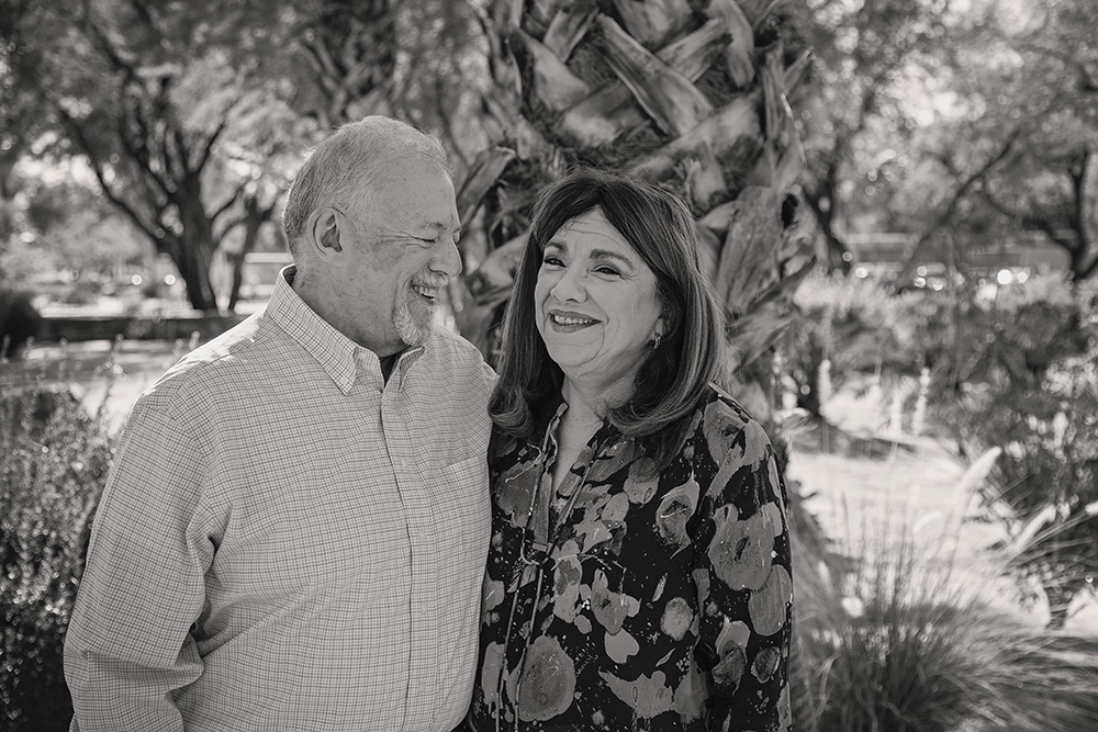 Grandparents standing together outdoors in Palm Springs, smiling warmly at each other in front of palm trees and desert foliage.