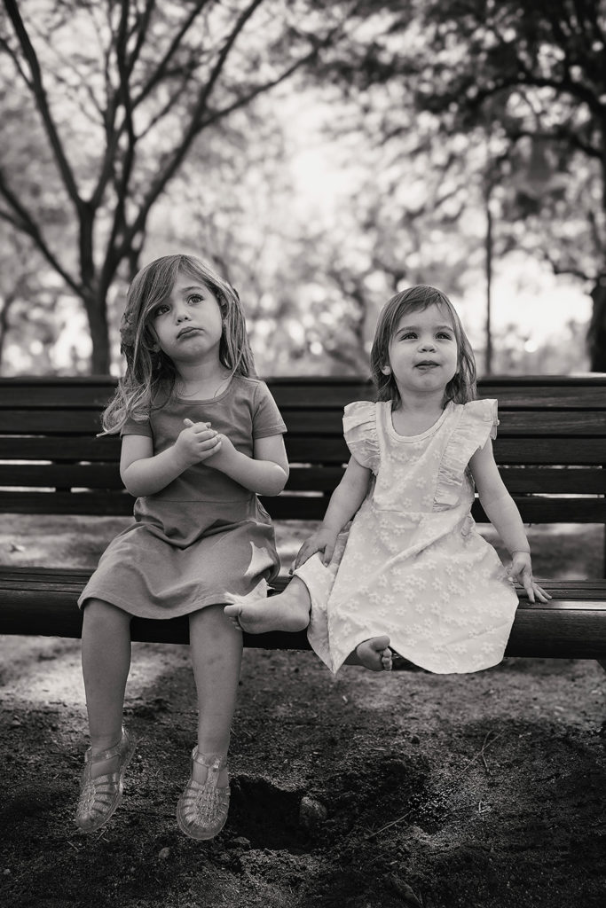 Two young girls sitting together on a park bench in Palm Springs, looking softly off camera in a calm, candid moment.