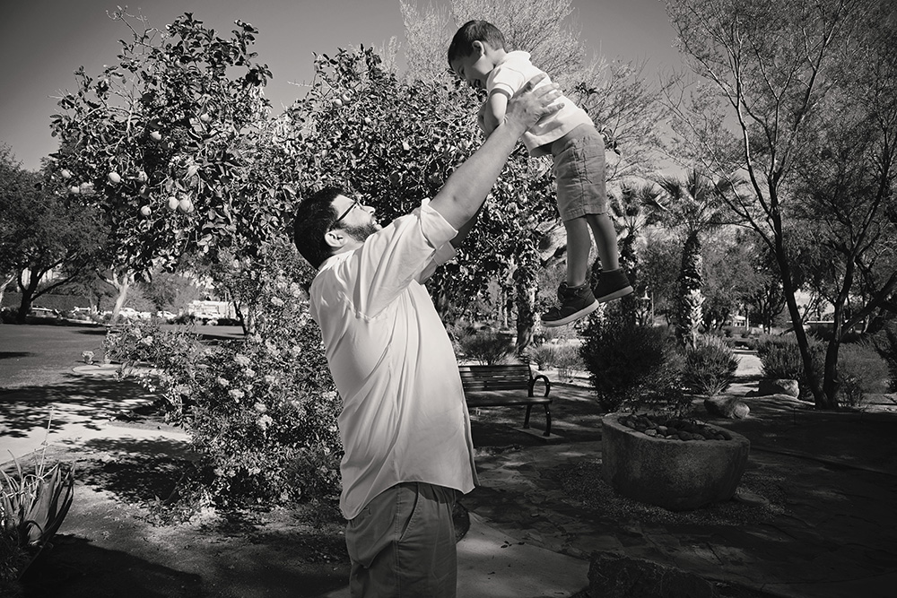 Parent lifting a young boy high into the air in a Palm Springs park, surrounded by citrus trees and desert foliage.