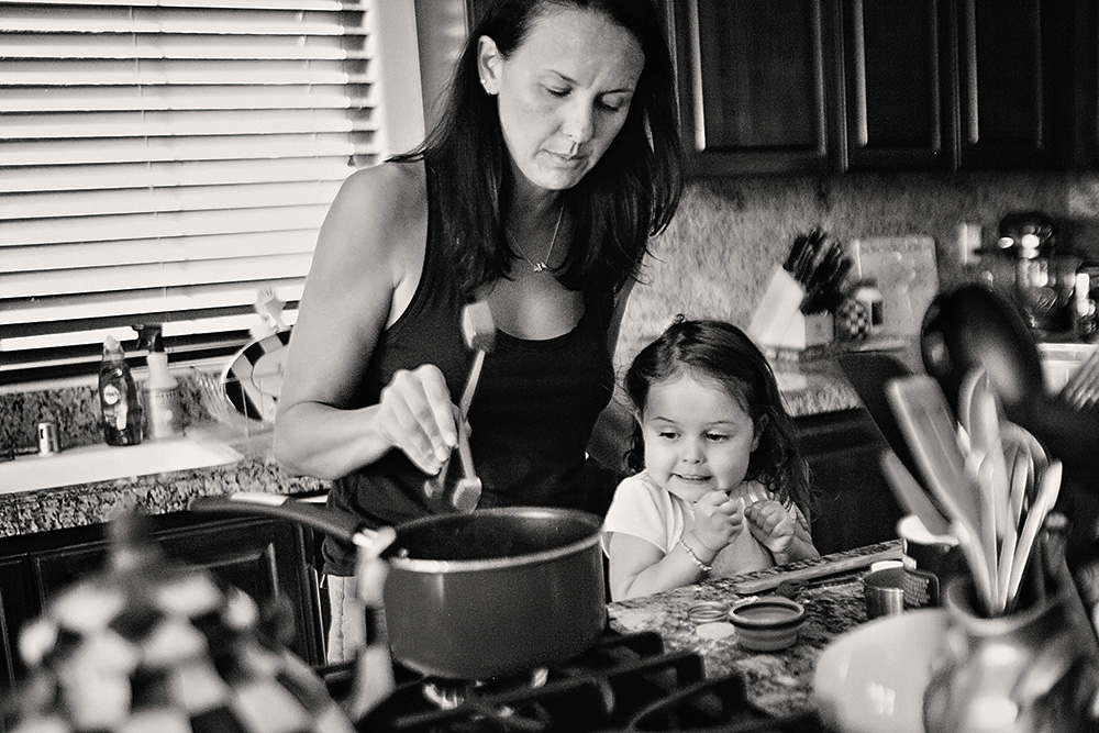 Mother cooking at the stove while her young daughter watches closely during a day in the life family photo session at home.