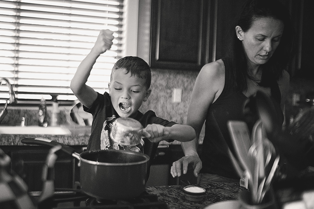 Young boy excitedly pouring ingredients into a pot while cooking with his mother during a day in the life family photo session.