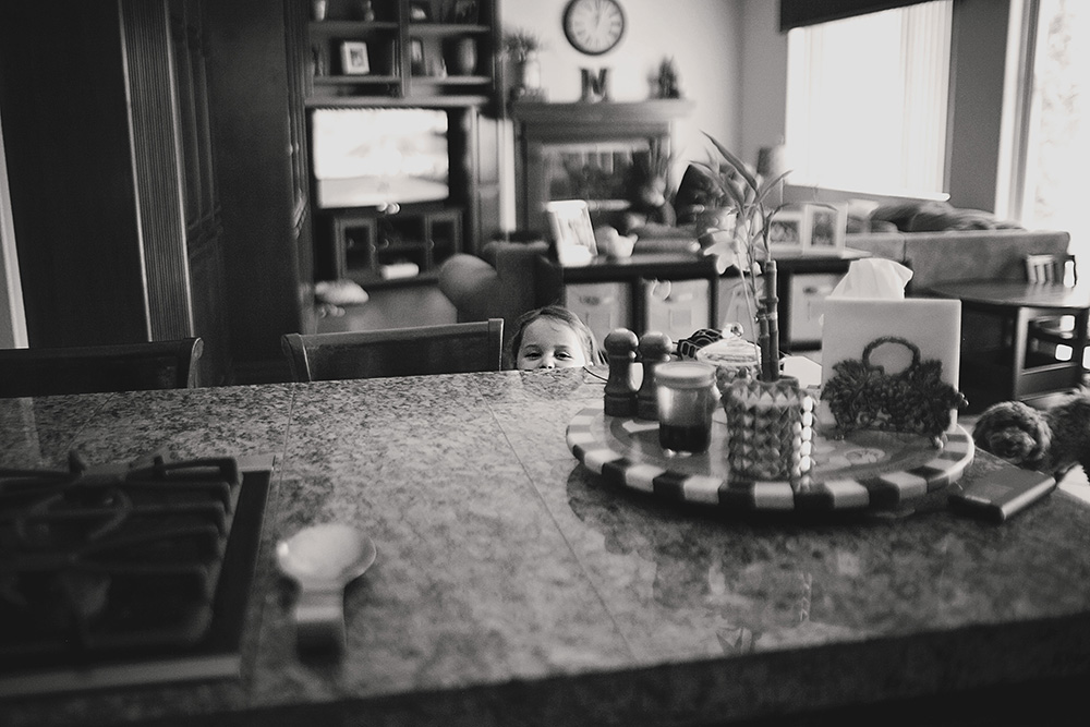 Toddler peeking over the kitchen counter during a candid day in the life family photo session at home.