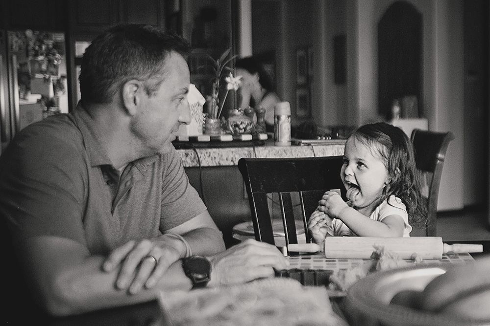 Father sitting at the table with his young daughter during a candid day in the life family photo session at home.