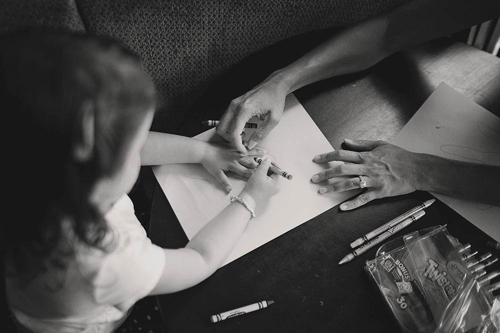 Toddler drawing with crayons alongside a parent at the table during a documentary day in the life family photo session.