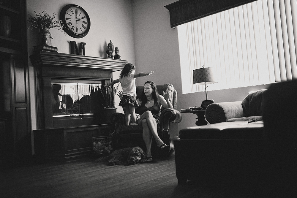 Mother sitting in a chair while her young daughter stands nearby in the living room during a day in the life family photo session.