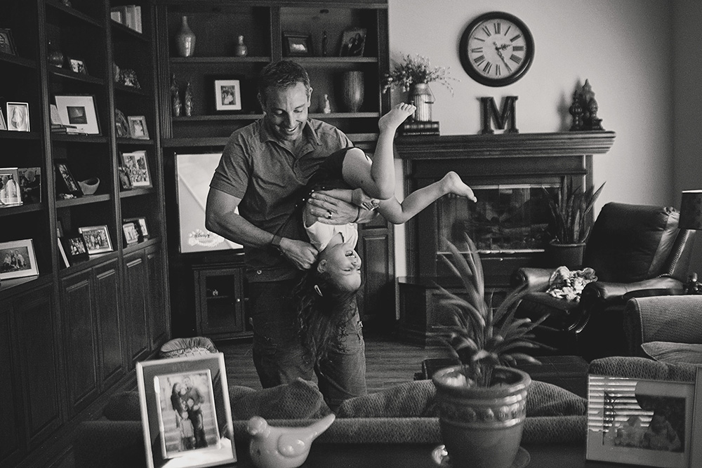 Father laughing while holding his daughter upside down in the living room during a candid day in the life family photo session.