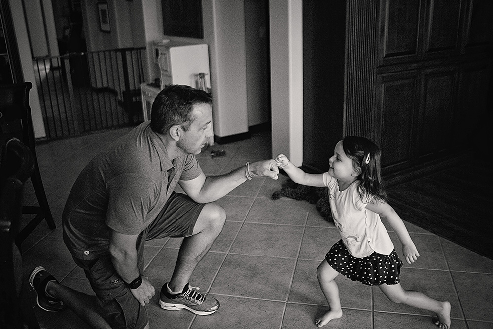 Father kneeling on the floor while his young daughter runs toward him during a candid day in the life family photo session.