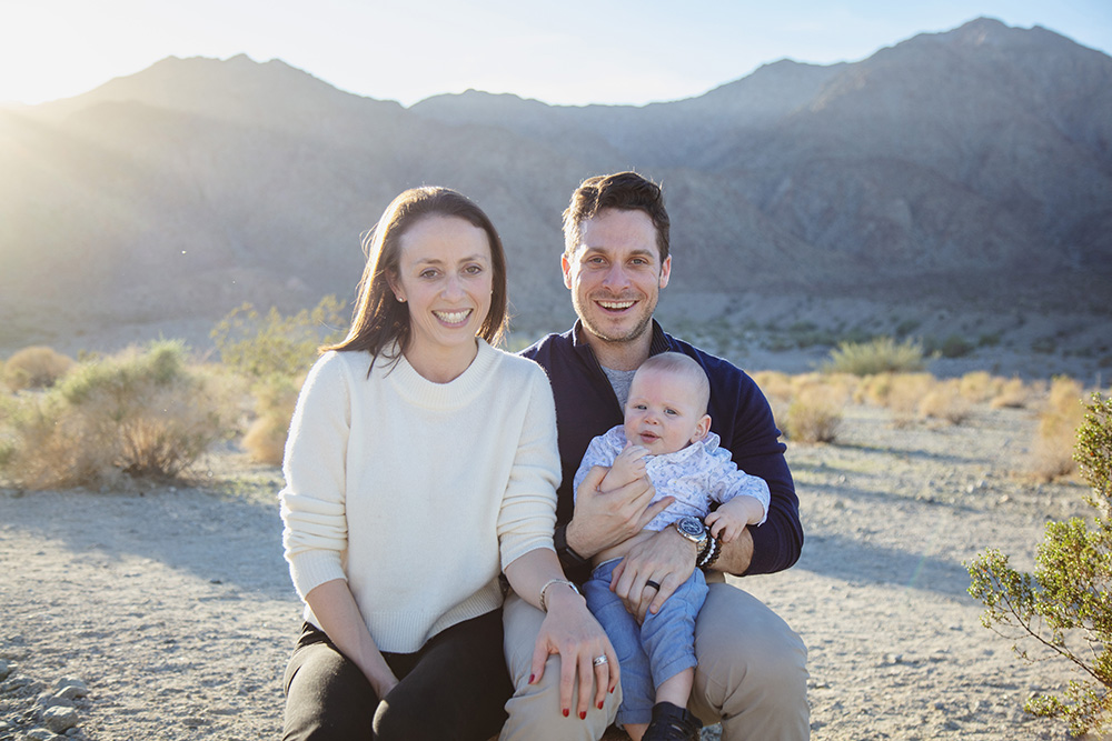 Family sitting together in La Quinta Cove with parents smiling and holding their baby, framed by desert plants and mountain views in soft evening light.